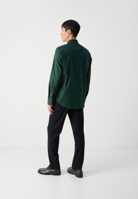 Camisa de manga larga verde oscuro con cuello puntiagudo, textura suave y puños abotonados, combinada con pantalones oscuros y zapatos negros.