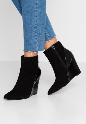 Personne portant un jean bleu effiloché et des bottines noires à talon compensé avec une fermeture éclair sur le côté, debout sur un sol blanc.