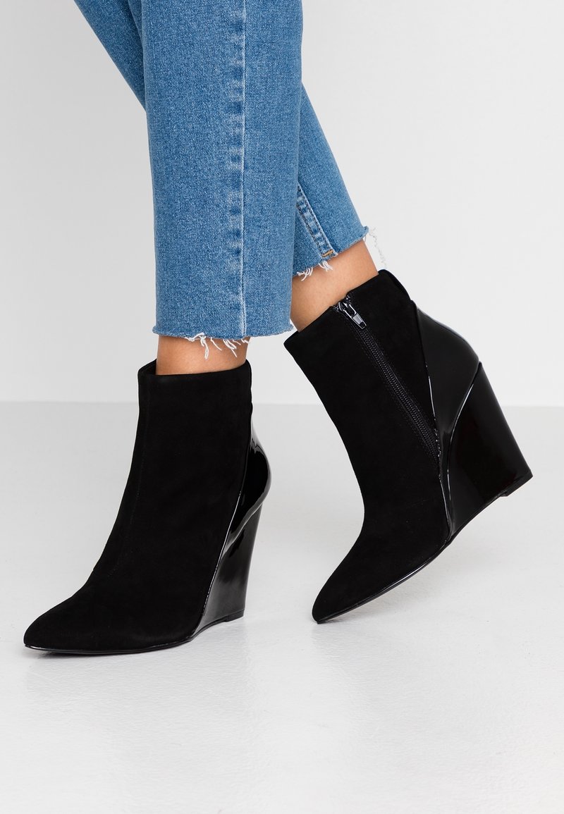 Personne portant un jean bleu effiloché et des bottines noires à talon compensé avec une fermeture éclair sur le côté, debout sur un sol blanc.
