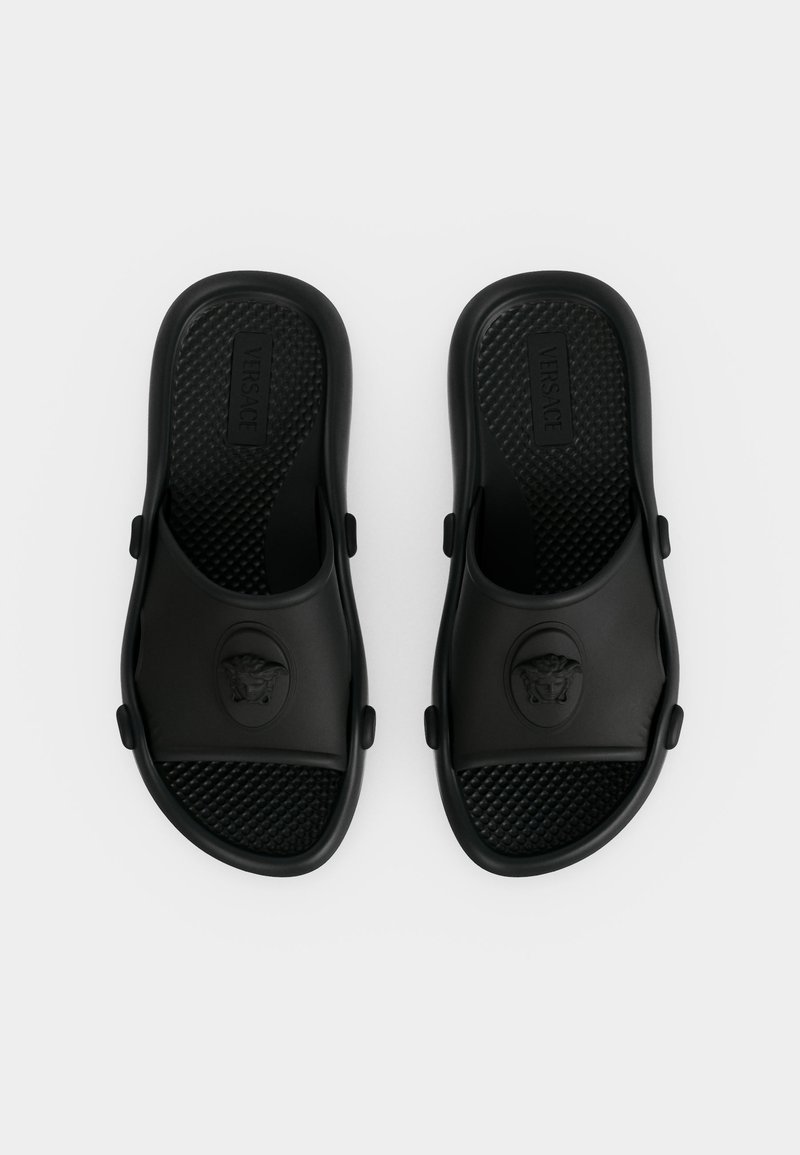 Coppia di sandali neri Versace slip-on con plantari testurizzati e emblema della testa di Medusa sulle fasce, mostrati dall'alto su sfondo bianco.