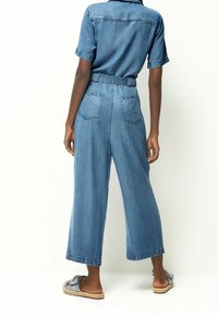 Džínový overal, světle modrý, krátké rukávy, široké nohavice, pas se zpevněním, dvě zadní kapsy, nošený s pletenými sandály na klínku.