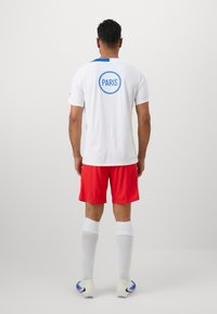 Hvid sportsjersey med blå accenter, der har "PARIS" i en blå cirkel på ryggen, parret med røde shorts og hvide sokker.