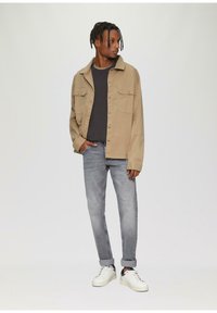 Beige shirtjack over een donkere trui, gecombineerd met lichtgrijze jeans en witte sneakers. Twee knoopzakken op de borst van de jack.