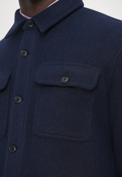 Sobrecamisa de mistura de lã azul-marinho com gola, botões pretos e dois bolsos de peito com abas, exibindo um acabamento de tecido texturizado e quente.