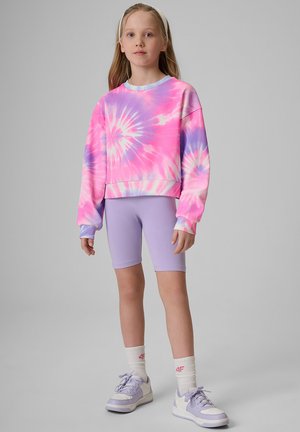 Młoda dziewczyna stoi, ubrana w różowo-fioletową bluzę tie-dye, jasnofioletowe spodenki, białe skarpetki i fioletowo-białe trampki.