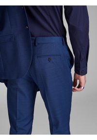 Blue tailored trousers with a textured finish, featuring a back pocket with a button closure. A navy shirt is partially visible.