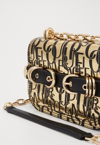 Borsa a tracolla metallica dorata con lettering nero, superficie liscia, doppi fibbie e tracolla nera removibile con accentuazione a catena.