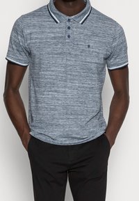 Chemise polo bleu marine avec un motif texturé, trois boutons et des bordures contrastantes au col et aux manches. Portée avec un pantalon noir.