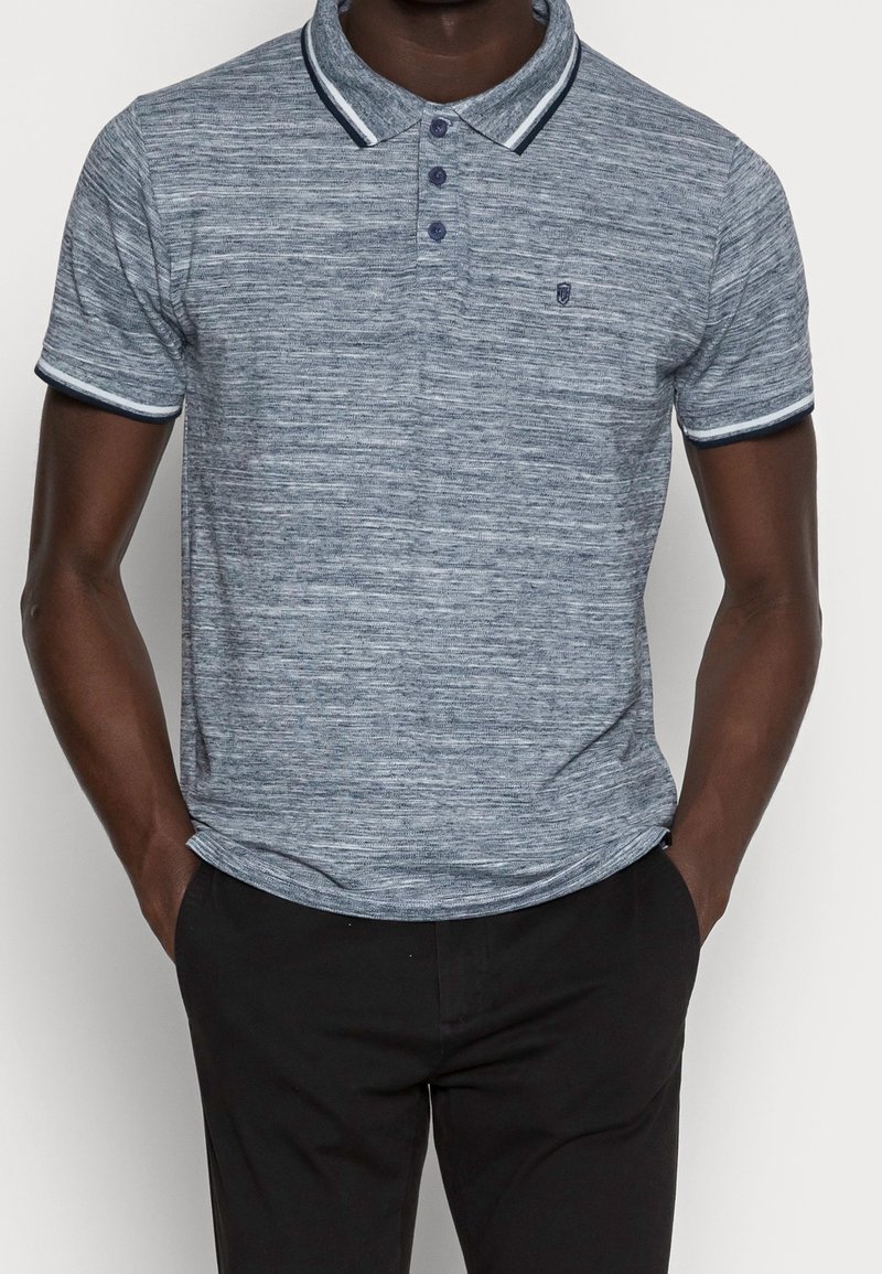 Chemise polo bleu marine avec un motif texturé, trois boutons et des bordures contrastantes au col et aux manches. Portée avec un pantalon noir.