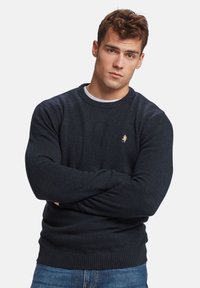 Suéter de punto azul oscuro con cuello redondo acanalado y puños. Presenta un pequeño logo dorado en el pecho. Lleva puesto sobre una camisa blanca.