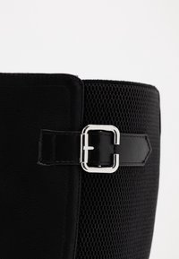 Acercamiento a una bota negra texturizada con un cierre de hebilla cuadrada plateada en una correa de cuero, sobre un fondo blanco.