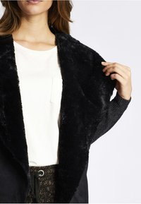 Black faux fur wrap with a soft texture, open front, and long sleeves. Worn over a white shirt, showcasing a relaxed fit.