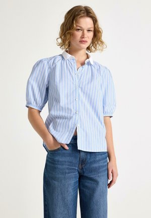 Kvinde iført en blå og hvidstribet kortærmet bluse med pufærmer og blå jeans, stående med den ene hånd i lommen.