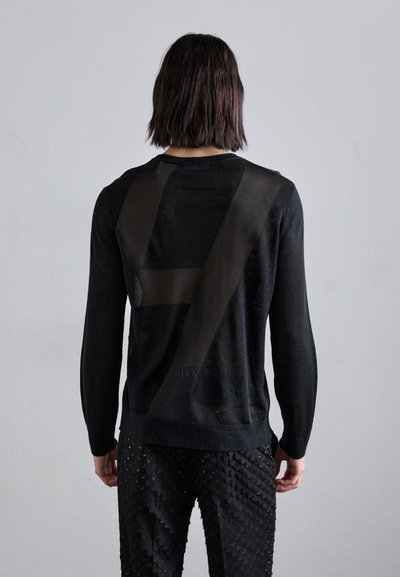 Sort strikket sweater med en semi-transparent ryg med et geometrisk mønster og lange ærmer, parret med teksturerede sorte bukser med skæl.
