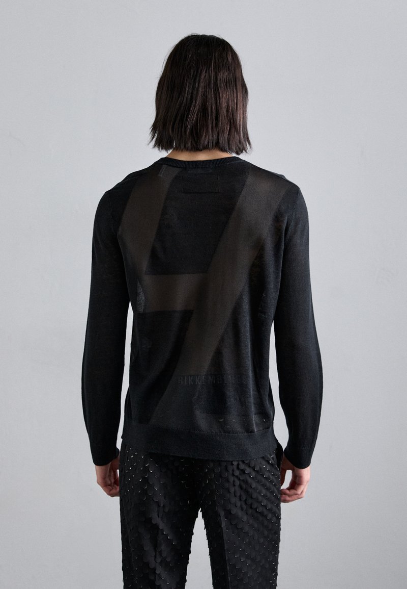Schwarzer Strickpullover mit einem semi-transparenten Rücken im geometrischen Muster und langen Ärmeln, kombiniert mit strukturierten schwarzen Hosen mit Schuppen.