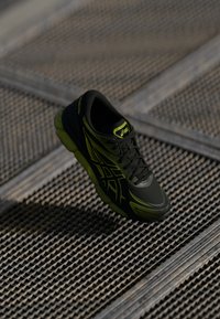 Črne atletske čevlje z zelenimi poudarki, z teksturirano zgornjimi deli, okroglo prstjo in vezalnim sprednjim delom, ki lebdi nad vzorčasto površino.