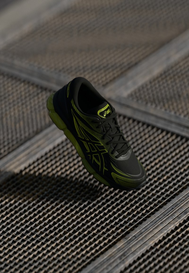 Črne atletske čevlje z zelenimi poudarki, z teksturirano zgornjimi deli, okroglo prstjo in vezalnim sprednjim delom, ki lebdi nad vzorčasto površino.