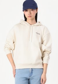 Felpa beige con tasca a marsupio e cappuccio con cordino, con un piccolo logo "new balance". Abbinata a jeans azzurri chiari. Cappellino blu navy indossato.