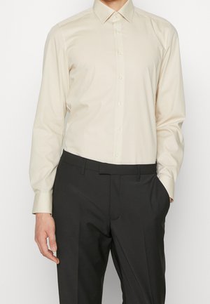 Mężczyzna noszący kremową koszulę z długim rękawem wpuszczoną w czarne formalne spodnie, z jedną ręką w kieszeni.