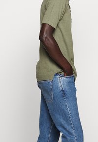 Olivově zelená polokošile s krátkým rukávem a žebrovaným límcem a spodním lemem, doplněná o světle modré džíny s pěti kapsami a vybledlým vzhledem.