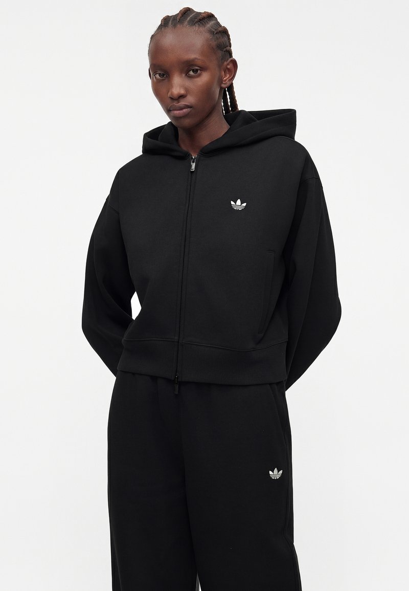 Personne portant un sweat à capuche zippé noir Adidas et un pantalon de survêtement Adidas noir assorti avec des détails de logo blancs, debout devant un fond uni.