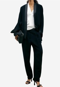 Veste noire de style kimono avec bordure en satin, associée à un chemisier blanc et un pantalon noir sur mesure. Présente une silhouette structurée et fluide.