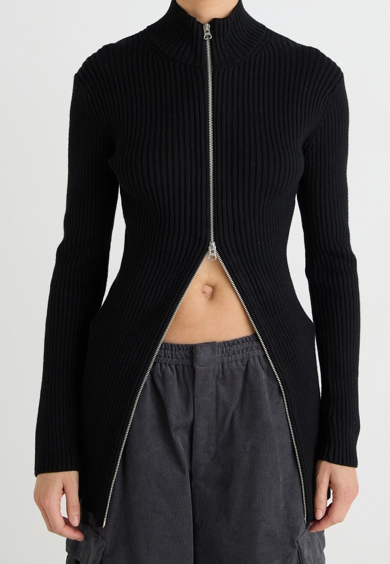 Pull noir en maille côtelée à col montant, doté d'une fermeture éclair à l'avant avec une fente à l'ourlet, une texture contrastante et une silhouette ajustée.
