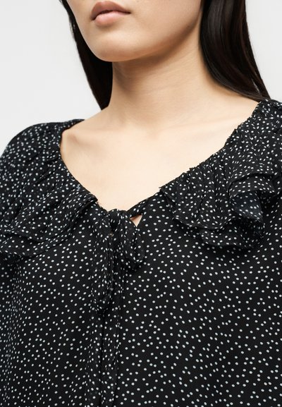 Γυναίκα που φοράει μαύρο μπλουζάκι με λευκές πουά, βολάν στο γιακά και κορδόνι μπροστά, εμφανίζεται από το λαιμό μέχρι το πάνω μέρος του στήθους.