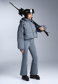 Grå pufferjacka med hög krage, i kombination med åtsittande grå skidbyxor. Svarta skidstövlar och skidor fulländade looken. Matta ytor på materialen.