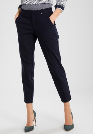 Vrouw die nauwsluitende, marineblauwe cropped broek draagt, met een patronen blouse ingestopt, handen in de zakken en groene hoge hakken aan.