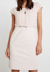 Robe rose clair avec des manches courtes style cape, dotée d'un décolleté en trou de serrure et d'un motif en losanges texturé. Comprend une ceinture fine nouée à la taille.