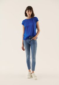 Blauw T-shirt met korte mouwen en een ontspannen pasvorm, gecombineerd met lichtblauwe skinny jeans en witte sneakers, tegen een neutrale achtergrond.