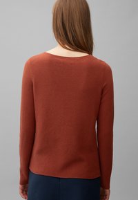 Pullover in Rostfarbe mit langen Ärmeln, weitem Ausschnitt und strukturiertem Strickmuster. Der Saum verläuft hinten leicht geschwungen.