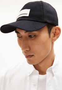 Donkerblauwe pet met een gebogen klep, voorzien van een witte rechthoekige patch met het label "ARMEDANGELS." Draagt een witte overhemden.