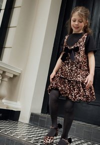 Sijajna obleka iz rožnatega zlata z sequini, s črno pentljo, z večplastnim naborkom na robu, kombinirana s črnimi nogavicami in leopardjim tiskom čevljev z pentljami.