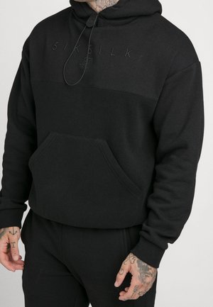 Mann trägt schwarzen Hoodie in Zweitonoptik und passende schwarze Hose, mit sichtbaren Tattoos an Händen und Hals, schaut nach vorne.