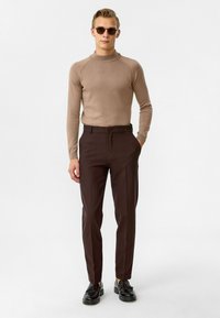 Bruine geribbelde trui met hoge hals, zwarte broek en zwarte schoenen. Model met ronde zonnebril, staande tegen een effen achtergrond.