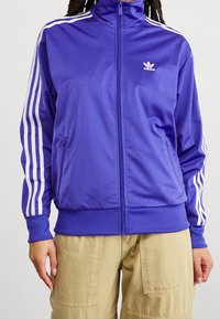 Lila satinträningsjacka med vit tre-stripesdesign på ärmarna, full blixtlås framtill och liten vit logotyp på bröstet.