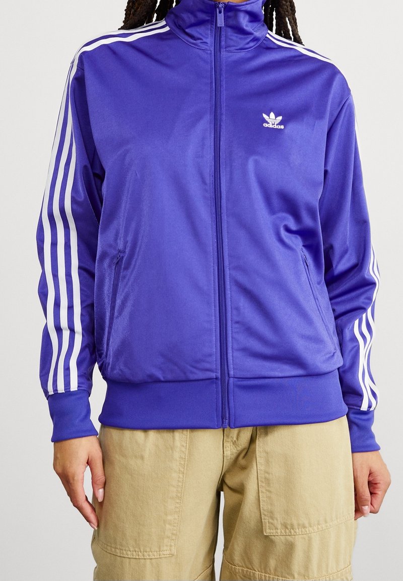 Lila satinträningsjacka med vit tre-stripesdesign på ärmarna, full blixtlås framtill och liten vit logotyp på bröstet.