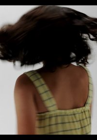 Kvinde med skulderlangt mørkt hår iført en gul ternet ærmeløs top, der vender hovedet, hvilket får håret til at virvle rundt.