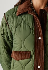 Green quilted jacket with a brown corduroy collar and trim. Features a button closure and a small pocket on the front.