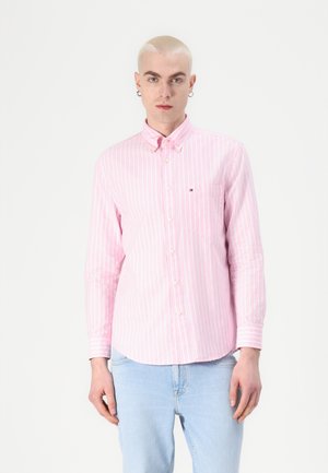 Ung person med bleket kort hår iført en rosa og hvit stripete skjorte med knapper og lyseblå jeans, sett forfra mot hvit bakgrunn.