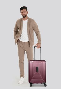 Valigia rigida di un profondo bordeaux con motivo a righe e una maniglia nera. Accompagnata da un uomo in un completo beige e scarpe bianche.