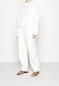 Weißer Strickpullover mit vertikaler Rippung, kombiniert mit locker sitzenden weißen Hosen und braunen Stiefeletten mit Muster.