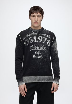 Jeune homme portant un pull noir à manches longues avec des lettres et motifs blancs délavés, debout devant un fond blanc uni.