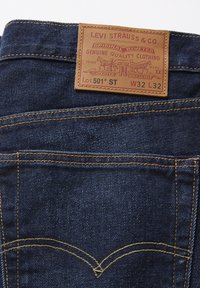Pantalones vaqueros de mezclilla de color azul oscuro, con una etiqueta de cuero que dice "Levi Strauss & Co." y costuras amarillas en los bolsillos traseros.