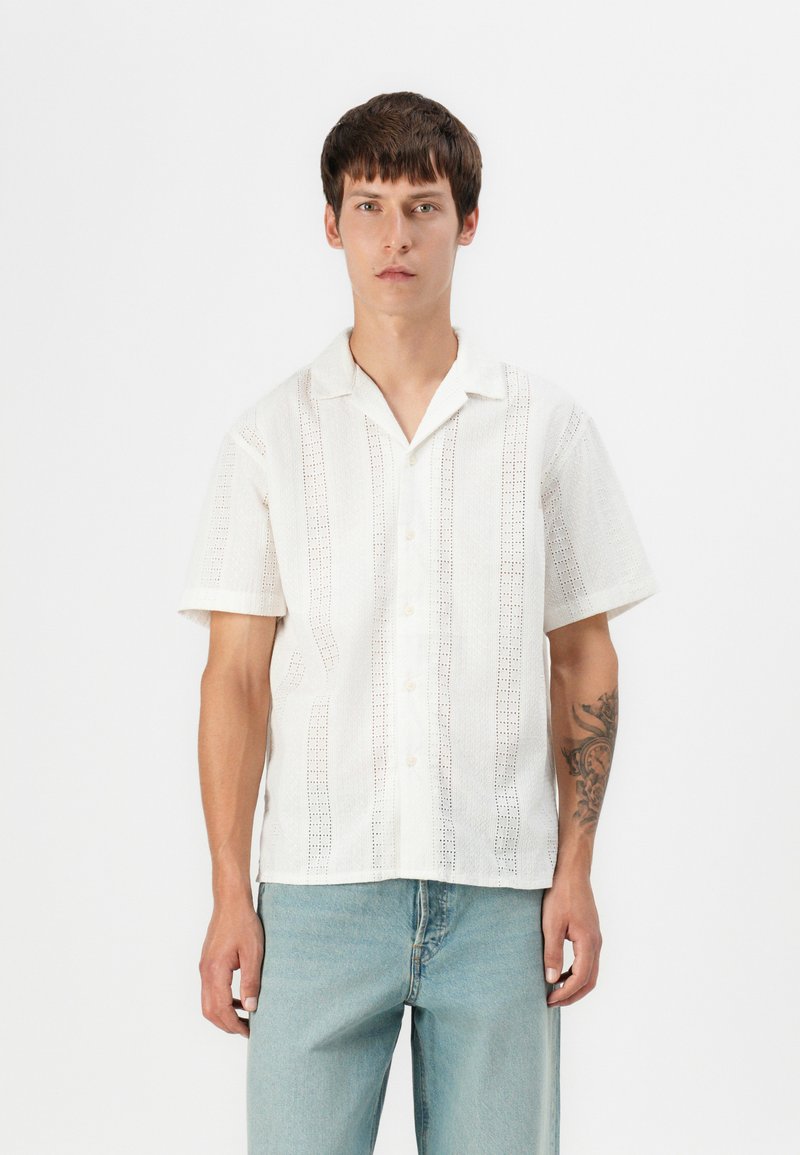 Camisa de manga corta, abotonada, de tejido blanco con patrones de corte vertical. Corte relajado, con cuello y bolsillos de parche.