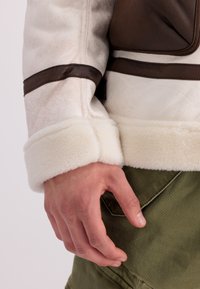 Bílá ovčí kožešinová bunda s hnědými koženými detaily. Rukáv je silný a měkký. Olivově zelené kalhoty viditelné v dolní části obrázku.