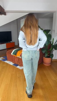 Osoba s dlhými hnedými vlasmi, ktorá má na sebe sivú mikinu a svetlomodré džínsy, stojí s rukami v bok v obývacej izbe s drevenou podlahou.