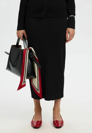 Set cardigan e gonna a coste nere, abbinato a una borsa a tracolla nera decorata con una sciarpa rossa e crema a contrasto, e scarpe basse rosse con punta quadrata.